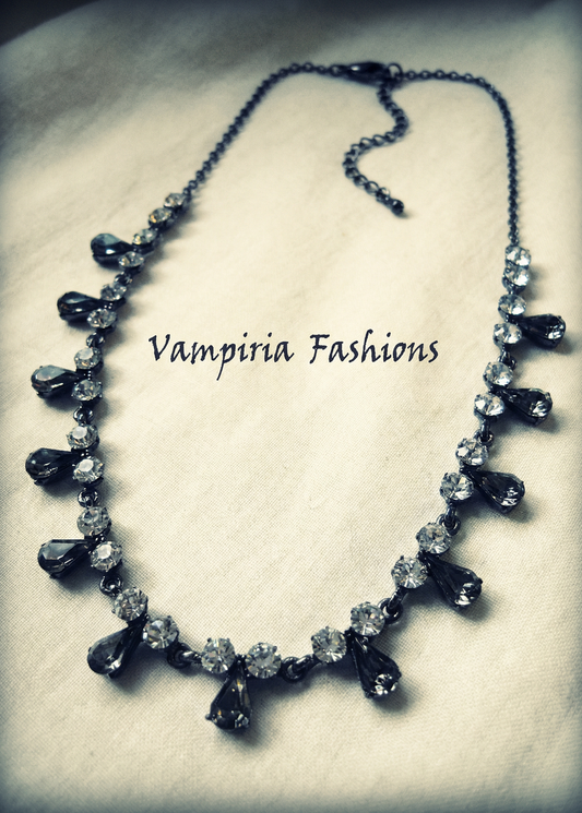 Schwarzes Strass-Collier mit dunklen Tropfenanhängern und Karabinerverschluss auf heller Stoffunterlage, mittig platziert.