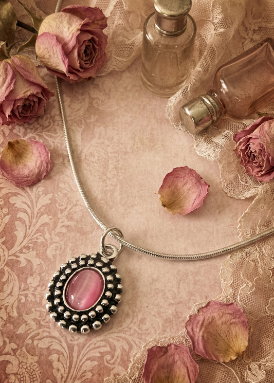 Silberfarbene Schlangenkette mit ovalem rosa Amulettanhänger auf rosa Untergrund mit getrockneten Rosen und Spitzenstoff.