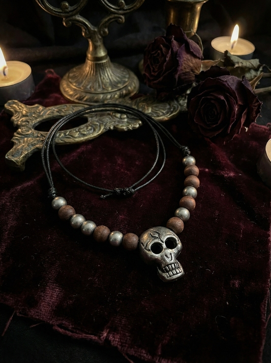 Halskette mit Totenkopf-Anhänger, Holzperlen und Metallperlen auf dunkelrotem Samtstoff neben Kerzen, Rosen und dekorativen Metallobjekten.