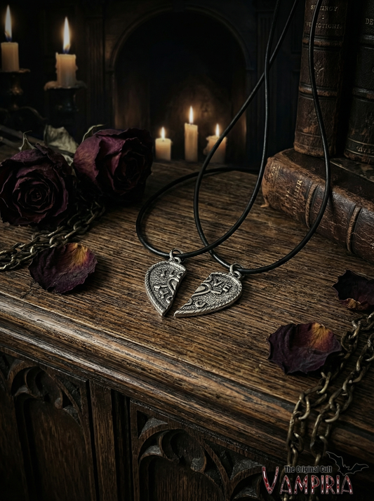 Zwei schwarze Kordelketten mit geteiltem Herzanhänger in Antik-Silberoptik auf Holzoberfläche vor Kerzen, dunklen Rosen, Kette und alten Büchern; unten rechts Schriftzug „Vampiria“.