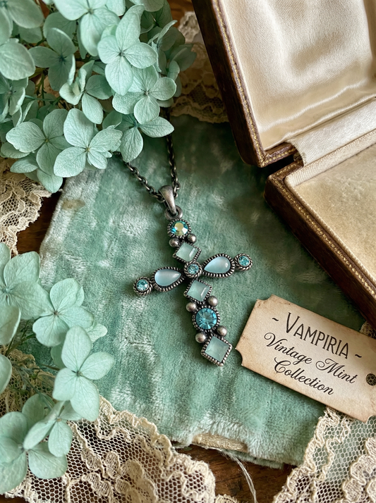 Kreuzanhänger im Antikdesign mit mintfarbenen und türkisfarbenen Steinen an dunkler Gliederkette auf mintgrünem Stoff neben hellen Hortensienblüten, Spitze, geöffnetem Etui und Karte „Vampiria Vintage Mint Collection“.