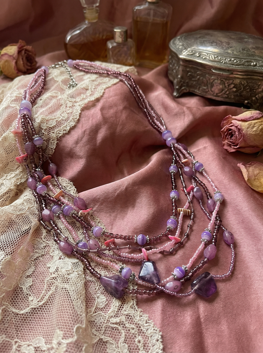 Mehrreihige Perlenkette in Rosa-, Flieder- und Lilatönen mit verschiedenen Perlen und lila Stein-Elementen auf rosafarbenem Satin neben Spitze, Glasflakons, Metallbox und getrockneten Rosen.