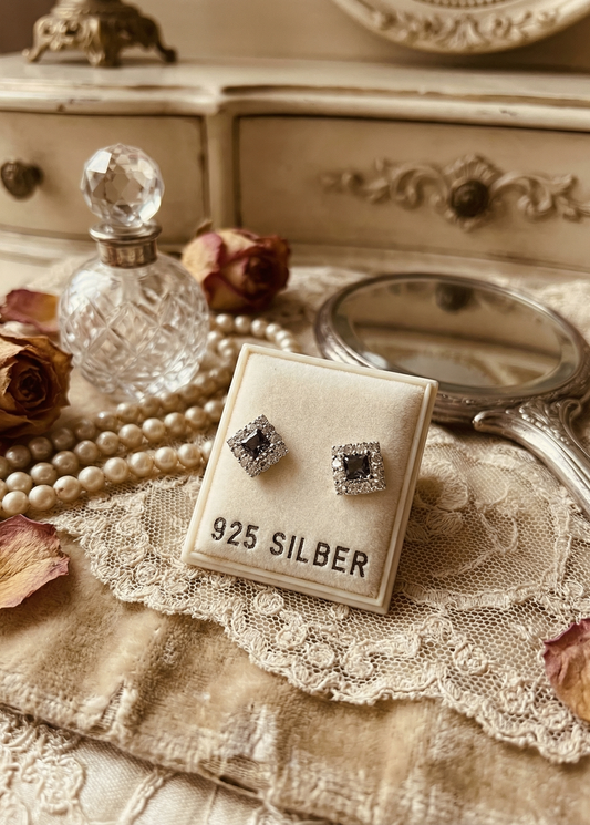 Zwei quadratische Mini Ohrstecker mit schwarzem Kristall und funkelndem Rand auf einem Display mit Aufschrift „925 SILBER“, dekoriert mit Spitze, Spiegel und Schmuckaccessoires.