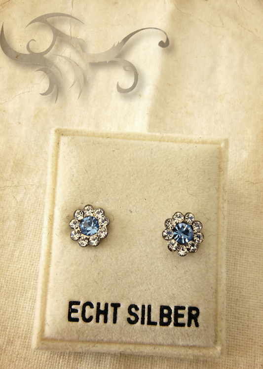 Zwei kleine Blumen-Ohrstecker mit hellblauem Stein auf einer hellen Karte mit Aufschrift „ECHT SILBER“ vor hellem Stoffhintergrund.