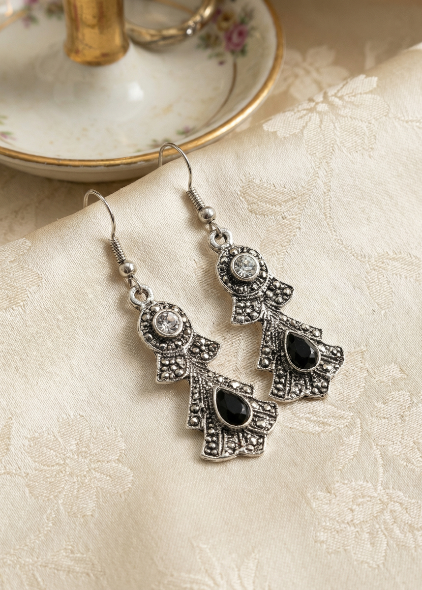 Zwei silberfarbene barocke Hängeohrringe mit schwarzen Tropfensteinen und klaren Strasssteinen auf hellem gemustertem Stoff neben Porzellanständer.