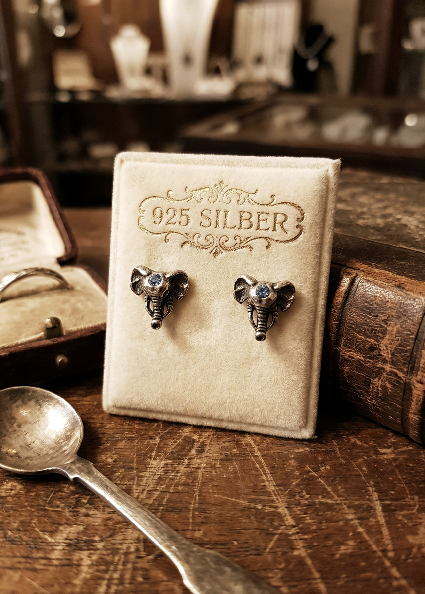 Zwei kleine Elefanten-Ohrstecker mit blauem Stein auf einem cremefarbenen Schmuckkärtchen mit Aufschrift „925 Silber“, fotografiert auf einem Holztisch mit Vintage-Dekoration.