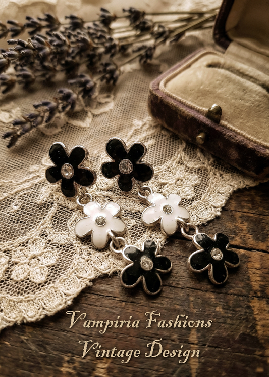 Schwarz-weiße Blumen-Ohrstecker im Vintage-Design mit drei hängenden Blütenelementen und kleinen klaren Steinen auf Holzuntergrund mit Spitze und Schriftzug.