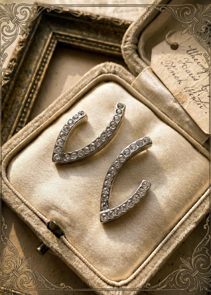 Zwei silberfarbene Ohrstecker in offener geschwungener Form mit klaren Strasssteinen auf einem cremefarbenen Schmuckkissen in einer Vintage-Schatulle, dekorativer Rahmen und Anhängerkarte im Hintergrund.