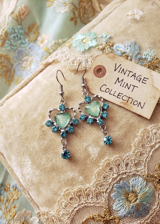 Zwei Vintage-Herzohrringe mit mintgrünem Herz und türkisblauen Kristallsteinen auf hellem Samtkissen neben Schild „VINTAGE MINT COLLECTION“ und Spitzenstoff.