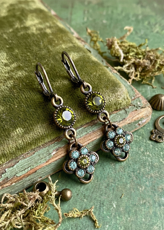 Zwei grüne Vintage-Ohrringe in Antik-Optik mit Hakenverschluss, rundem olivgrünem Stein und blumenförmigem Anhänger auf grünem Buch und Stoff.