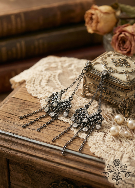 Zwei antikfarbene Vintage-Ohrringe mit filigranen Ornamenten, Strass und langen Kettenfransen liegen auf einem Holztisch neben einer verzierten Schmuckdose und Perlen.