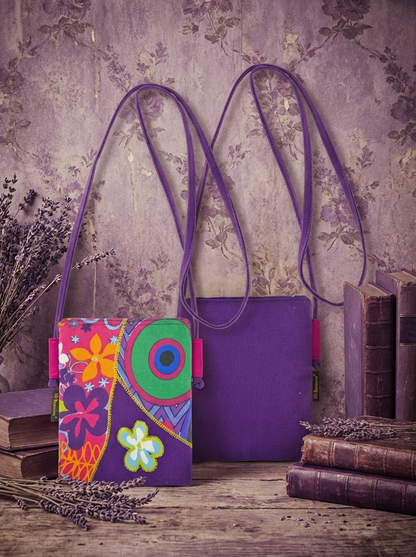 Zwei kompakte Umhängetaschen aus Baumwolle mit violettem Riemen, vorne Patchwork-Front mit bunten Blumen- und Kreismustern, hinten uni violette Rückseite, vor violetter Tapetenwand mit Büchern und Lavendelzweigen.