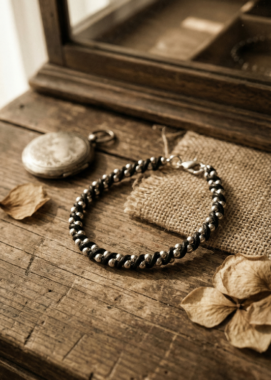 Schwarzes Armband mit silberfarbenen Perlen auf rustikalem Holztisch neben Taschenuhr und getrockneten Blättern.