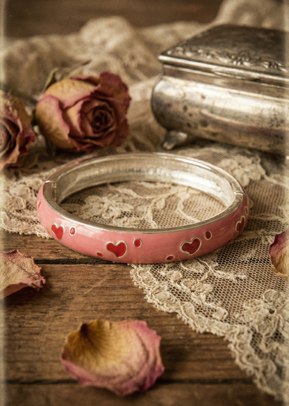 Rosa Armspange mit Herzmotiven auf Holztisch mit Spitze und getrockneten Rosen.