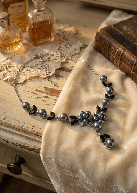 Dunkles Vintage Collier mit grauen Perlen und schwarzen Steinen liegt auf hellem Stoff neben Parfümflakons und Buch.