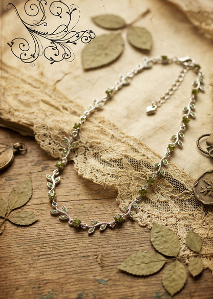 Silberfarbenes Blatt-Collier mit grünen Kristallen auf Holzuntergrund mit Spitze und getrockneten Blättern dekoriert.