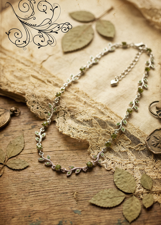 Silberfarbenes Blatt-Collier mit grünen Kristallen auf Holzuntergrund mit Spitze und getrockneten Blättern dekoriert.