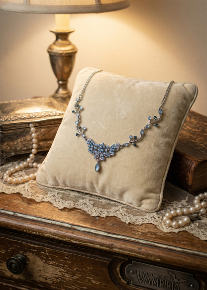 Filigranes silberfarbenes Collier mit blauen Kristallsteinen auf hellem Samtkissen neben Perlenkette und Vintage-Dekoration.