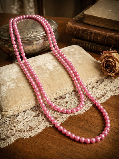 Lange pinke Perlenkette ohne Verschluss auf heller Samtunterlage mit Spitze, Holztisch, Buch und getrockneter Rose.