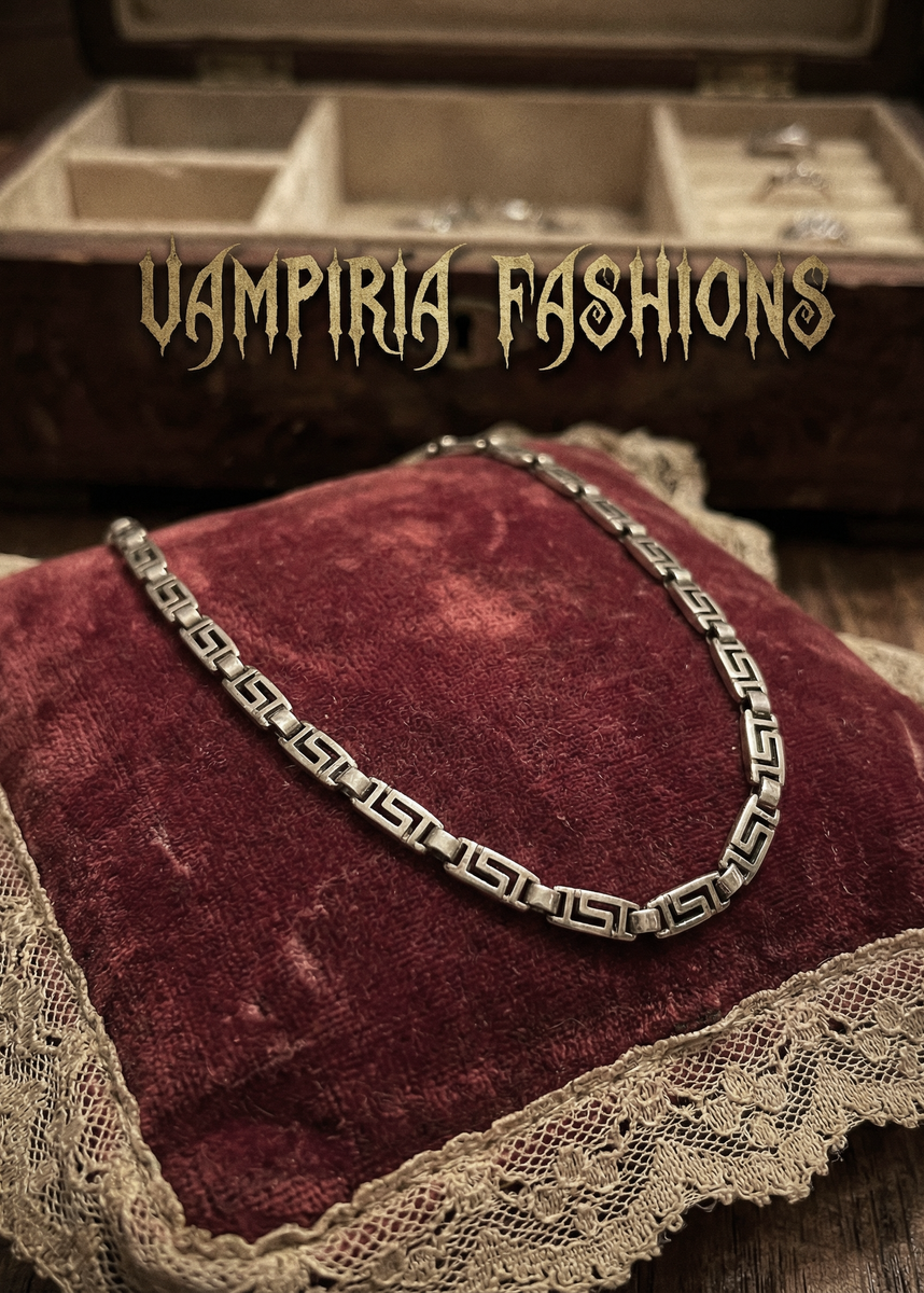 Silberfarbenes Metallcollier mit geometrischem Greek-Key-Muster auf bordeauxfarbenem Samtkissen, im Hintergrund Schmuckkästchen und Bücher.