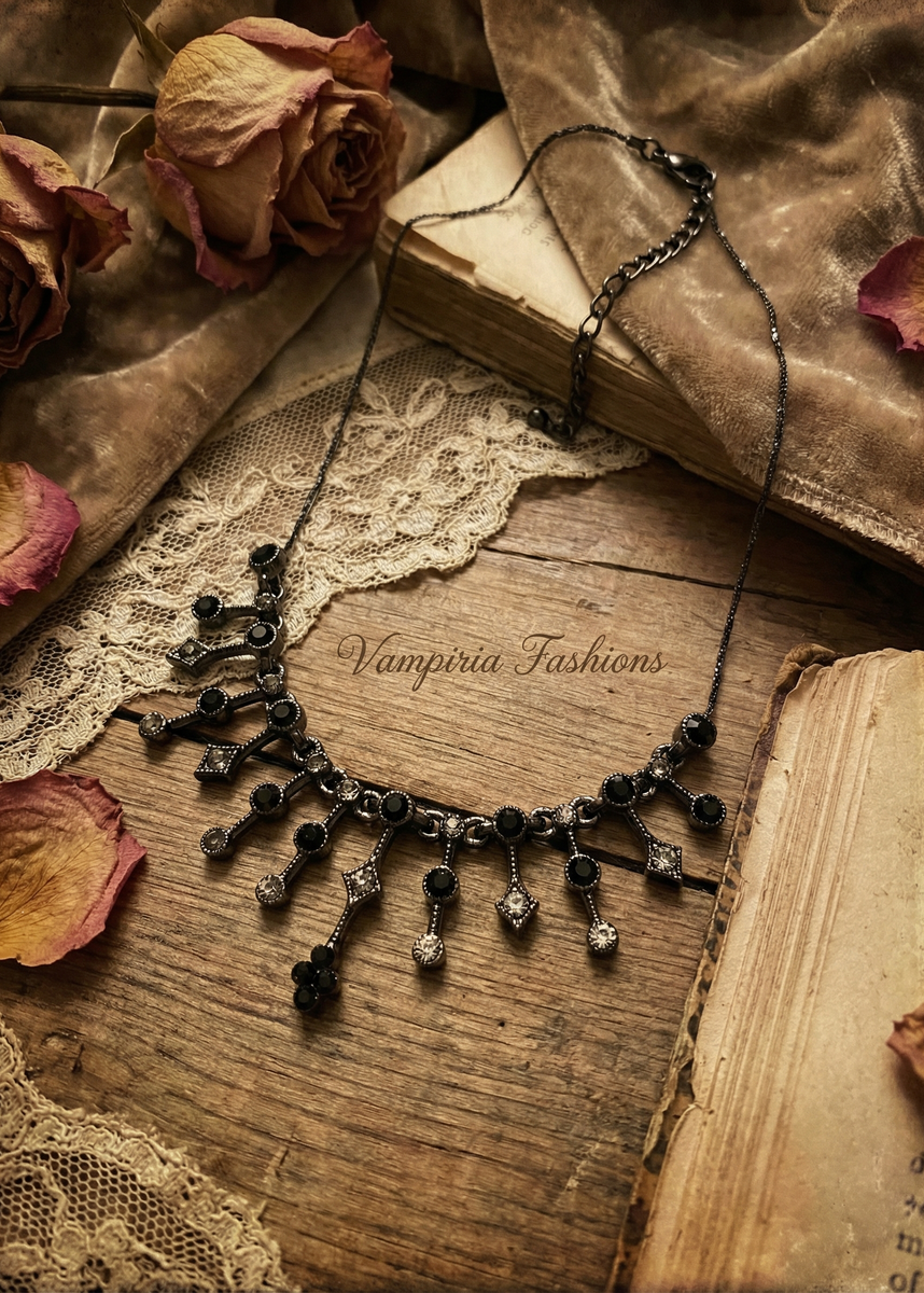 Schwarzes Gothic-Collier mit Strasssteinen und filigranen Anhängern auf Holzuntergrund zwischen alten Büchern, Spitze und getrockneten Rosen.