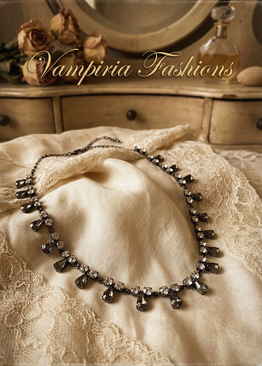 Antik wirkendes schwarzes Strass-Collier mit klaren Strasssteinen und dunklen tropfenförmigen Anhängern auf heller Stoff- und Spitzenunterlage vor Vintage-Schminktisch.
