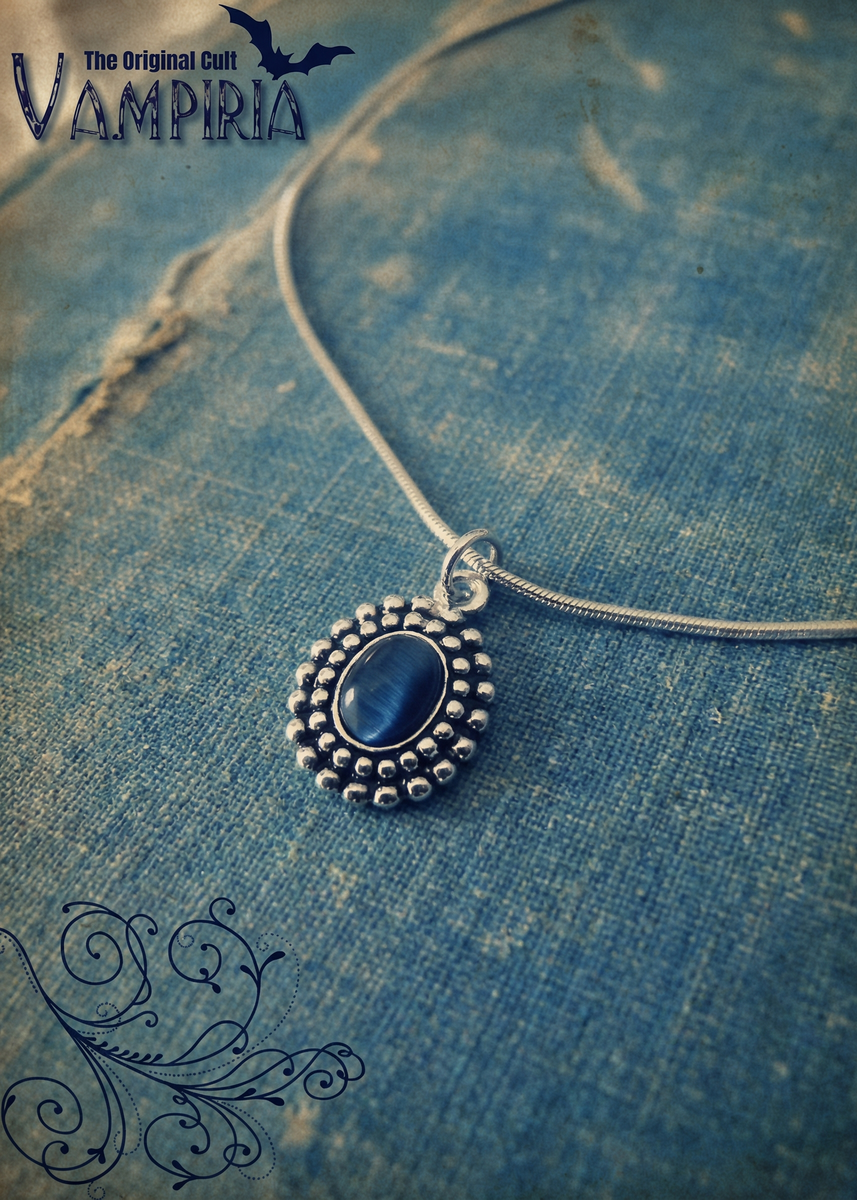 Silberfarbene Kette mit ovalem blauem Amulett auf blauem Stoff, Vampiria-Branding im Bild.