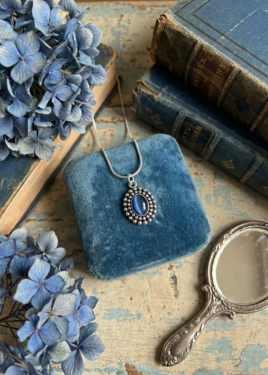 Silberfarbene Schlangenkette mit blauem ovalem Anhänger auf blauem Samtkissen zwischen blauen Blumen und alten Büchern.