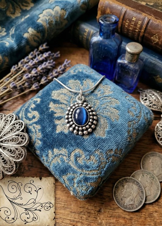 Nahaufnahme eines ovalen blauen Amuletts mit silberfarbenem Perlenrahmen auf blau gemustertem Kissen, mit Deko aus Fläschchen und Münzen.