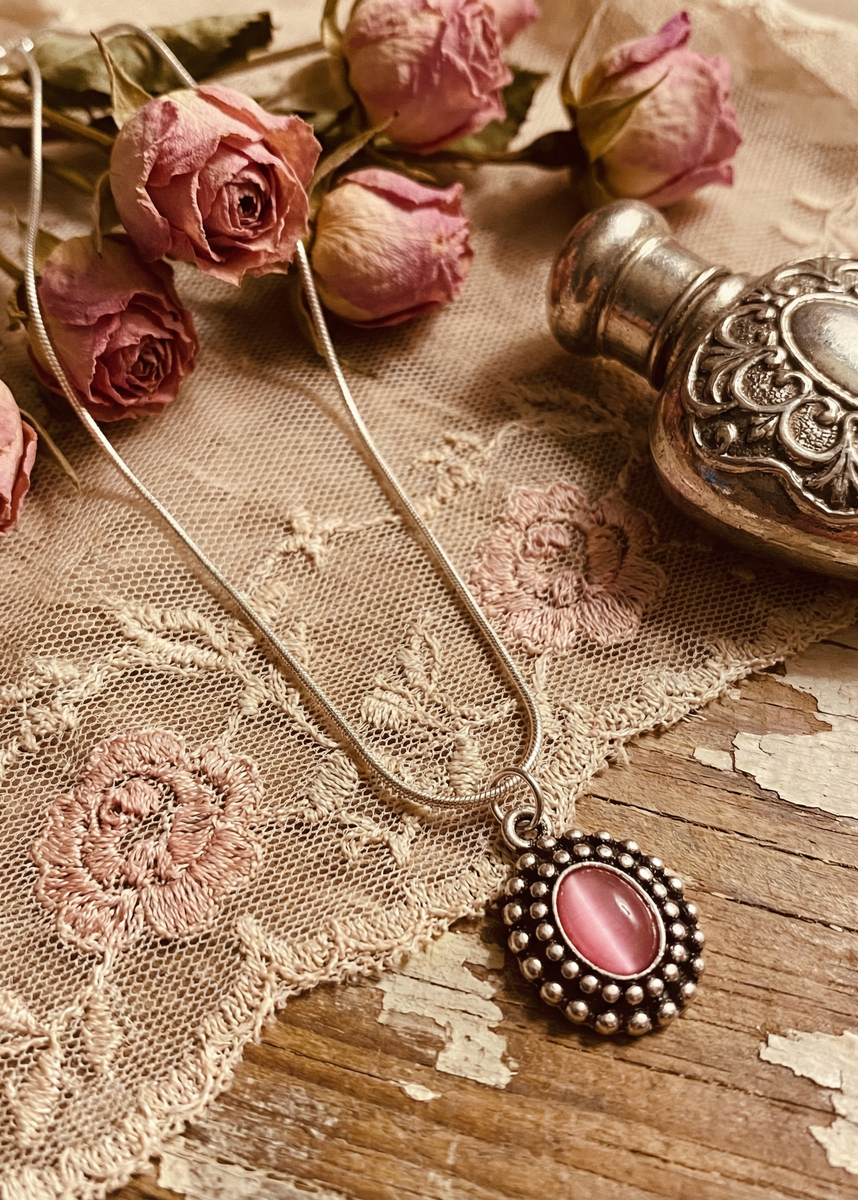 Silberfarbene Schlangenkette mit rosa ovalem Amulettanhänger auf Spitze und Holz neben Rosen und silberfarbener Dose.