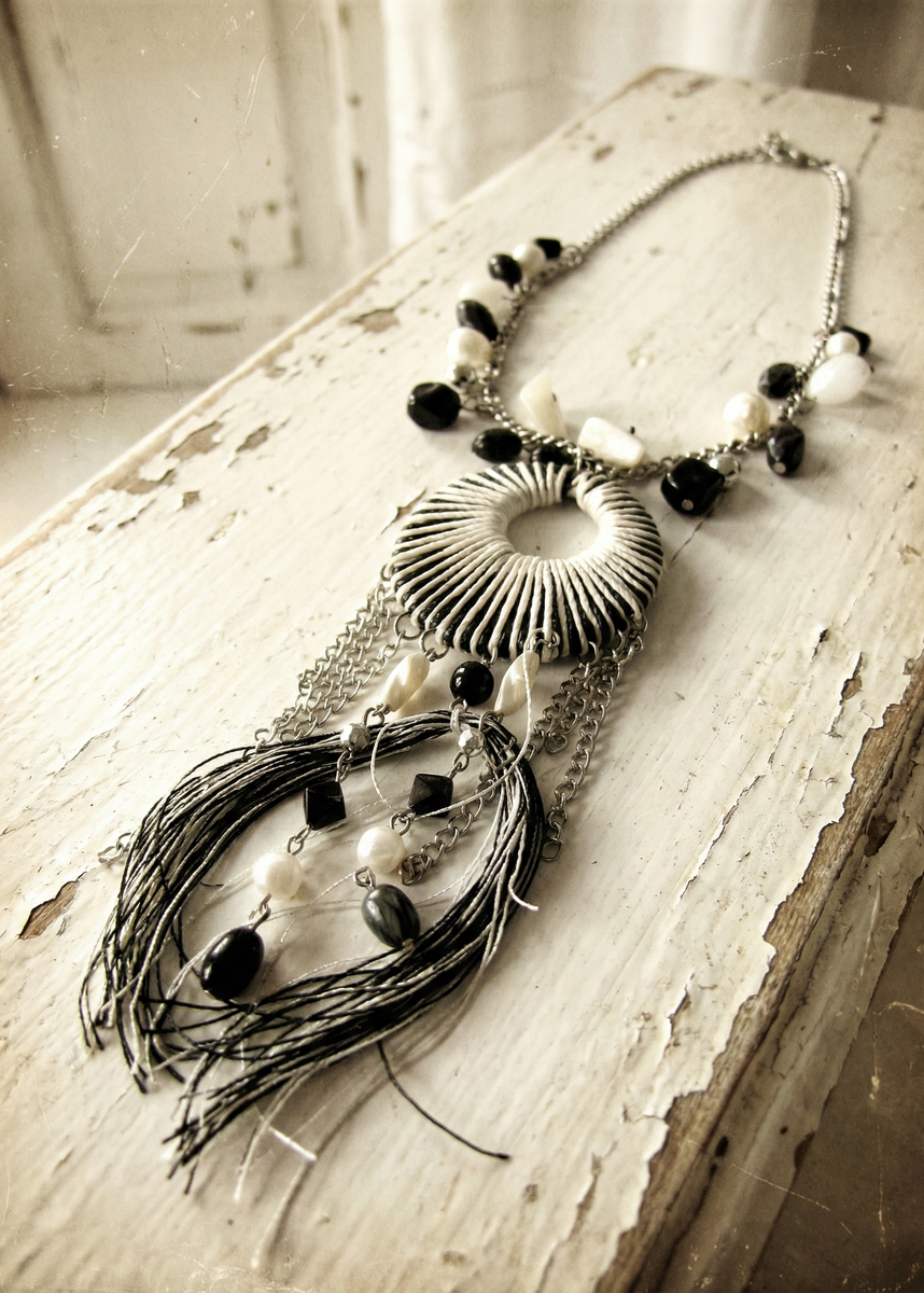 Boho-Halsschmuck in Schwarz-Weiß mit silberfarbener Kette, großem rundem Anhänger und langen Fransen auf weißer Holzoberfläche.