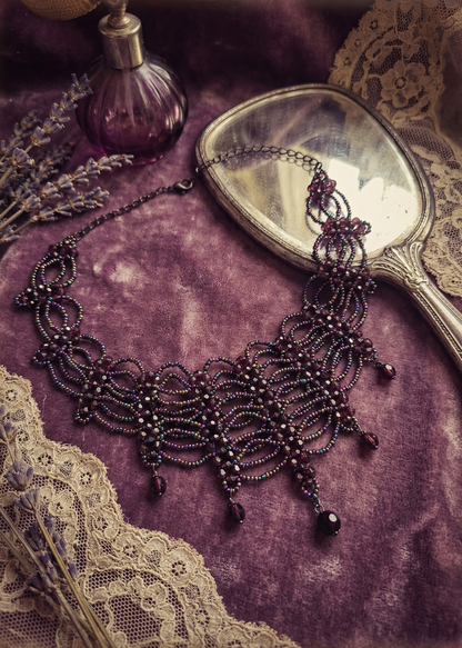 Violettes Gothic-Collier mit filigraner Perlenstruktur und Tropfenanhängern auf violettem Stoff neben Spiegel und Parfumflakon.