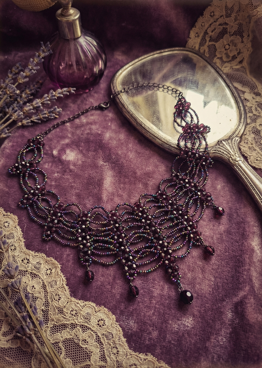 Violettes Gothic-Collier mit filigraner Perlenstruktur und Tropfenanhängern auf violettem Stoff neben Spiegel und Parfumflakon.