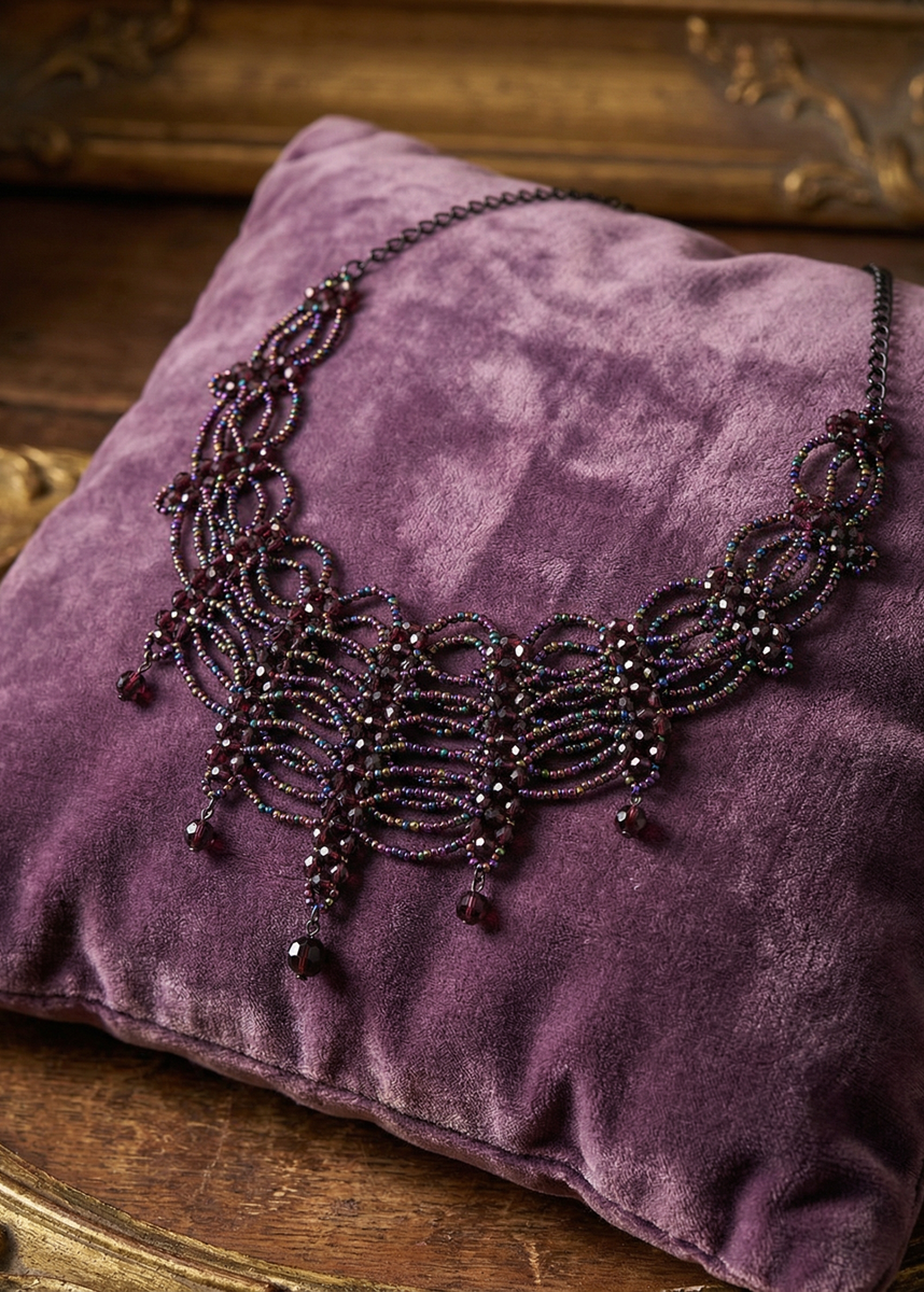 Violettes Gothic-Collier mit geschwungenem Perlenmuster und dunklen Tropfenanhängern auf einem violetten Samtkissen.