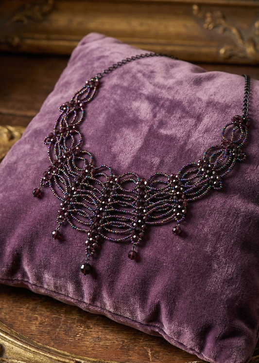 Violettes Gothic-Collier mit geschwungenem Perlenmuster und dunklen Tropfenanhängern auf einem violetten Samtkissen.