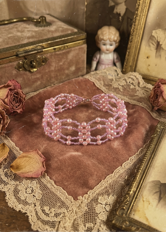 Rosa Perlen-Choker mit mehrreihigem Muster auf rosa Samtkissen, umgeben von Spitze, Trockenrosen und Vintage-Dekoration.