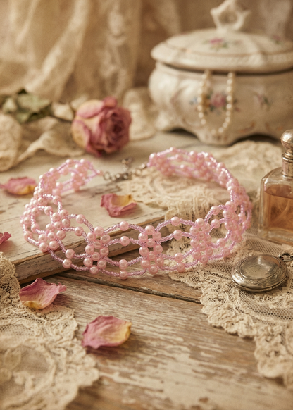 Rosa Lolita-Perlen-Choker mit floralem Perlenmuster auf Holzuntergrund mit Spitze, Glasflakon und getrockneten Rosenblättern.