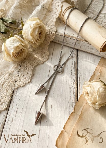 Silberfarbene Pendel-Kette mit Ring und zwei Kegelanhängern auf weißer Holzoberfläche neben getrockneten Rosen und Spitze.