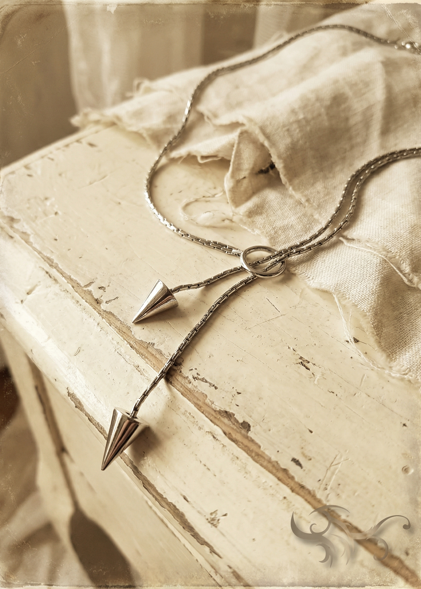 Silberfarbene Pendel-Kette mit Ring und zwei Kegelanhängern auf heller Kommode mit Stoff im Vintage-Stil.