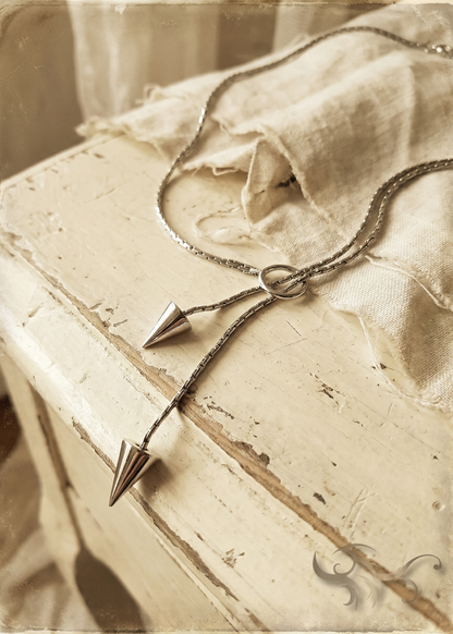 Silberfarbene Pendel-Kette mit Ring und zwei Kegelanhängern auf heller Kommode mit Stoff im Vintage-Stil.