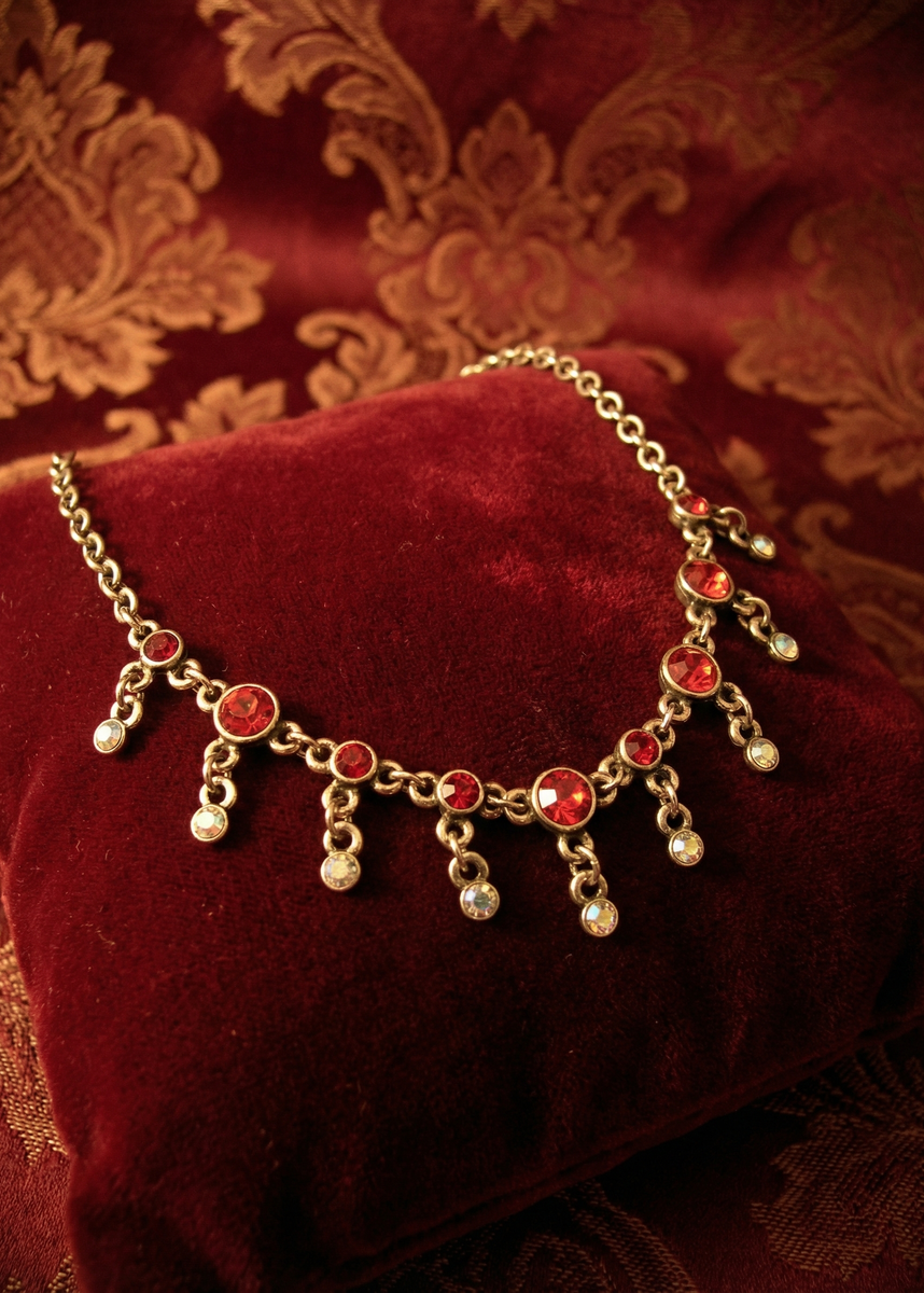 Rotes Collier mit roten und klaren Schmucksteinen auf rotem Samtkissen vor rotem ornamentalen Hintergrund.