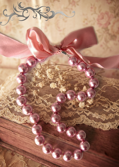 Rosa Perlenkette mit großen glänzenden Perlen und breitem rosa Satinband auf Spitze und Holzuntergrund.