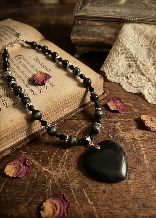 Schwarze Perlenkette mit großem schwarzem Herzanhänger auf einem alten Buch, rundherum liegen einzelne Rosenblätter und Spitze im Hintergrund