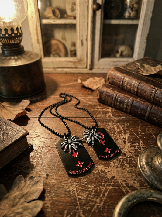 Zwei Gothic Partnerketten mit Spinnen-Dogtags auf einem Holztisch, dekoriert mit Vintage-Büchern und Lampe im Hintergrund.
