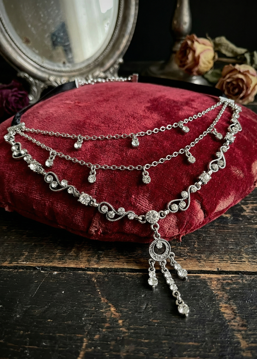 Silberfarbenes Vintage-Collier mit geschwungenen Ornamenten, klaren Steinen und langem Anhänger, liegend auf einem roten Samtkissen auf dunklem Holztisch, Spiegel und Rosen im Hintergrund.