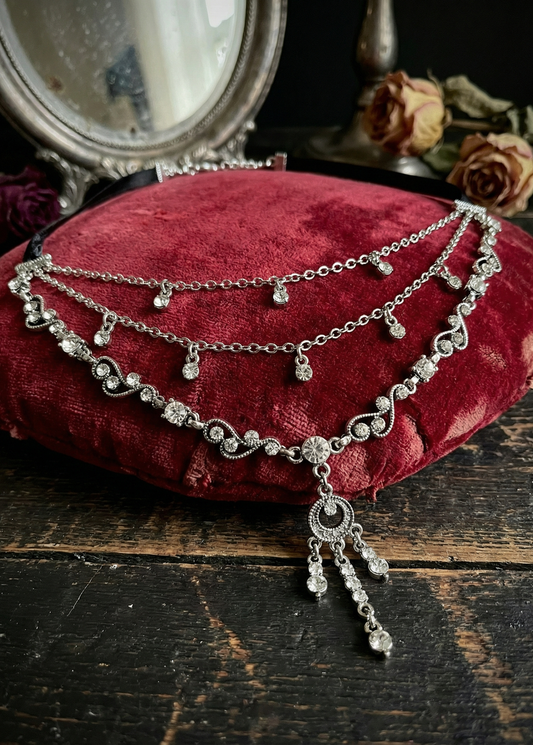 Silberfarbenes Vintage-Collier mit geschwungenen Ornamenten, klaren Steinen und langem Anhänger, liegend auf einem roten Samtkissen auf dunklem Holztisch, Spiegel und Rosen im Hintergrund.
