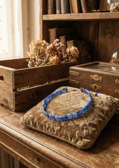 Blaues Perlen-Choker liegt auf einem Kissen vor Holzbox und Trockenblumen im Hintergrund.