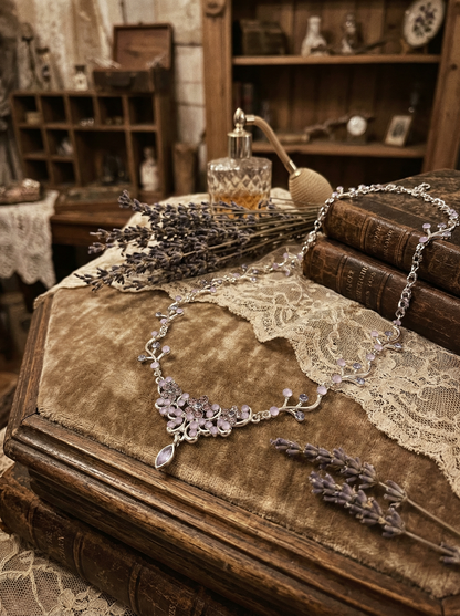 Florales Vintage-Collier mit lilafarbenen und roséfarbenen Steinen auf alten Büchern neben Lavendel, Spitze und Parfümflakon in dekorativer Retro-Umgebung.