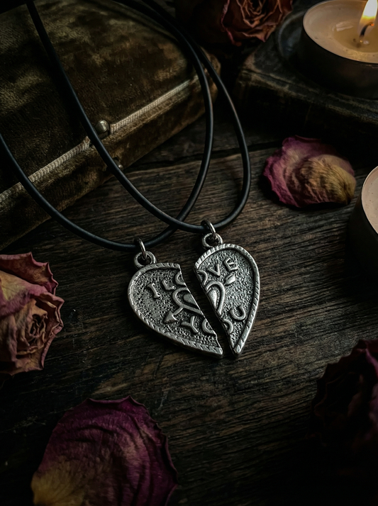 Nahaufnahme von zwei schwarzen Kordelketten mit geteiltem Herzanhänger und „LOVE YOU“-Schriftzug auf dunkler Holzoberfläche zwischen Rosenblättern, Buch und Kerzen.