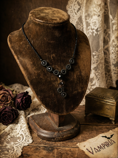 Schwarzes Y-Collier mit runden Hämatit-Ringen und schwarzen Perlen auf brauner Schmuckbüste vor Spitze, getrockneten Rosen und kleiner Metallbox; unten rechts Karte mit Schriftzug „Vampiria“.